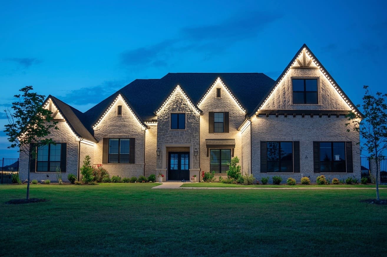 Large brick two-story home at dusk with warm white permanent lighting tracing every gable and eave of the roofline