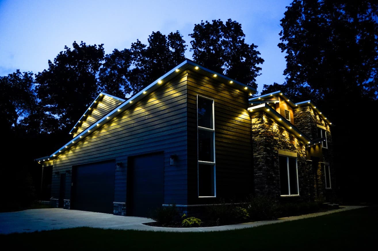 Side view of a dark-sided home and three-car garage at dusk with warm white permanent lights evenly spaced along the garage roofline