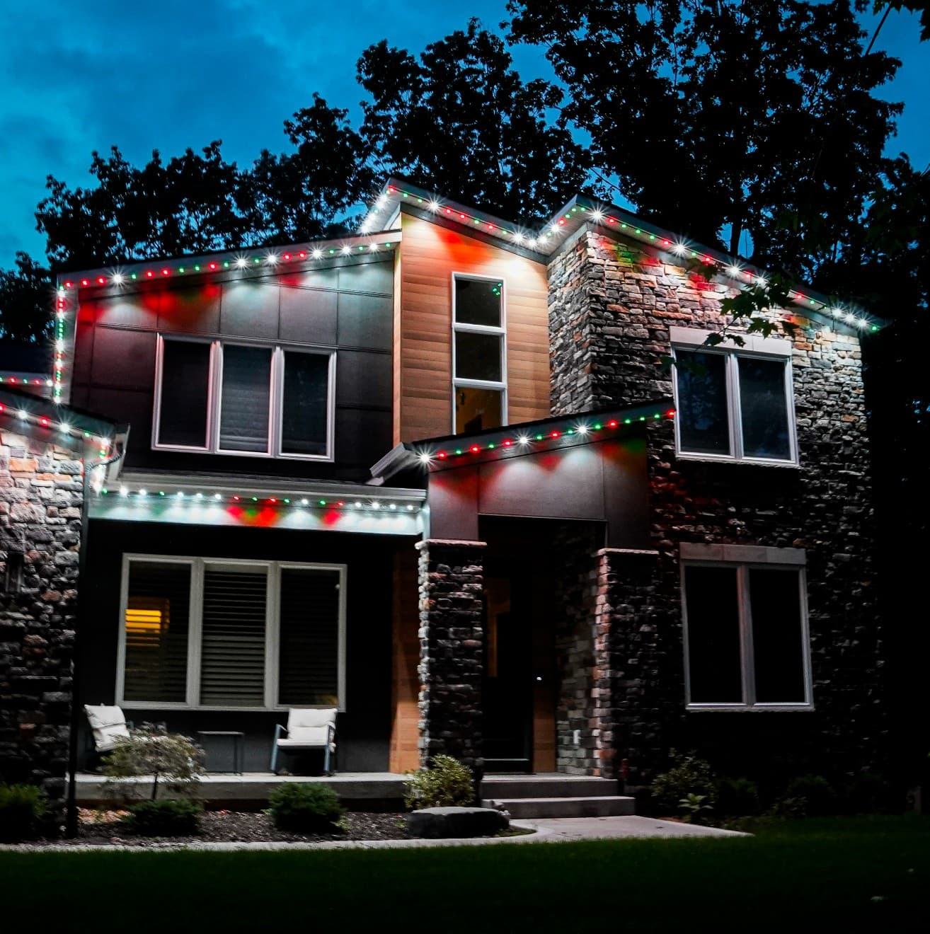 Modern two-story home with stone and wood siding, roofline lit in a repeating red, green, and white holiday pattern at night