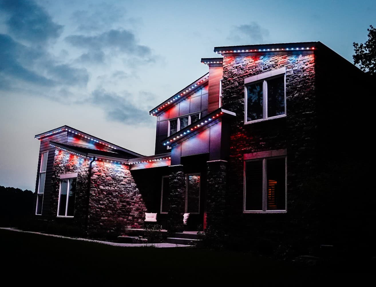 Modern stone home with angled rooflines at dusk, each roofline lit in red, white, and blue lights under a cloudy sky