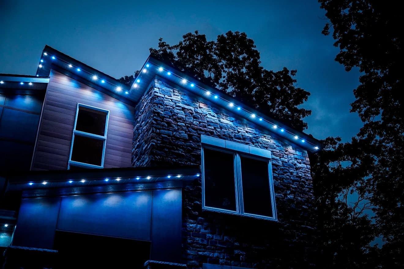 Stone and wood-sided home at dusk with blue and white permanent lights along its gables and garage roofline
