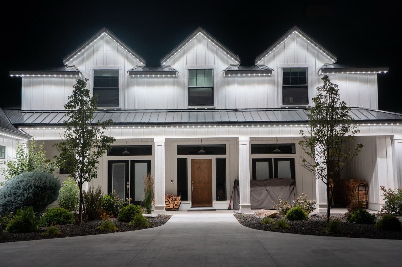 White modern farmhouse with three dormers at night, roofline and porch lit in bright white permanent lighting that washes the whole front of the home